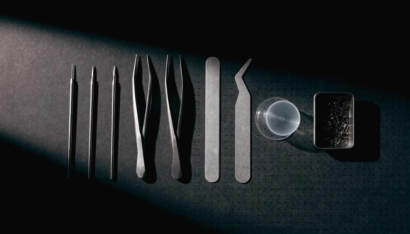 Precision repair tools laid out in a row on a dark bench mat: drivers, tweezers, pry tools, a suction cup and a tray of screws.